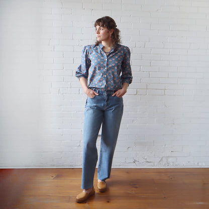 Person wearing a patterned shirt and blue jeans standing against a white brick wall.