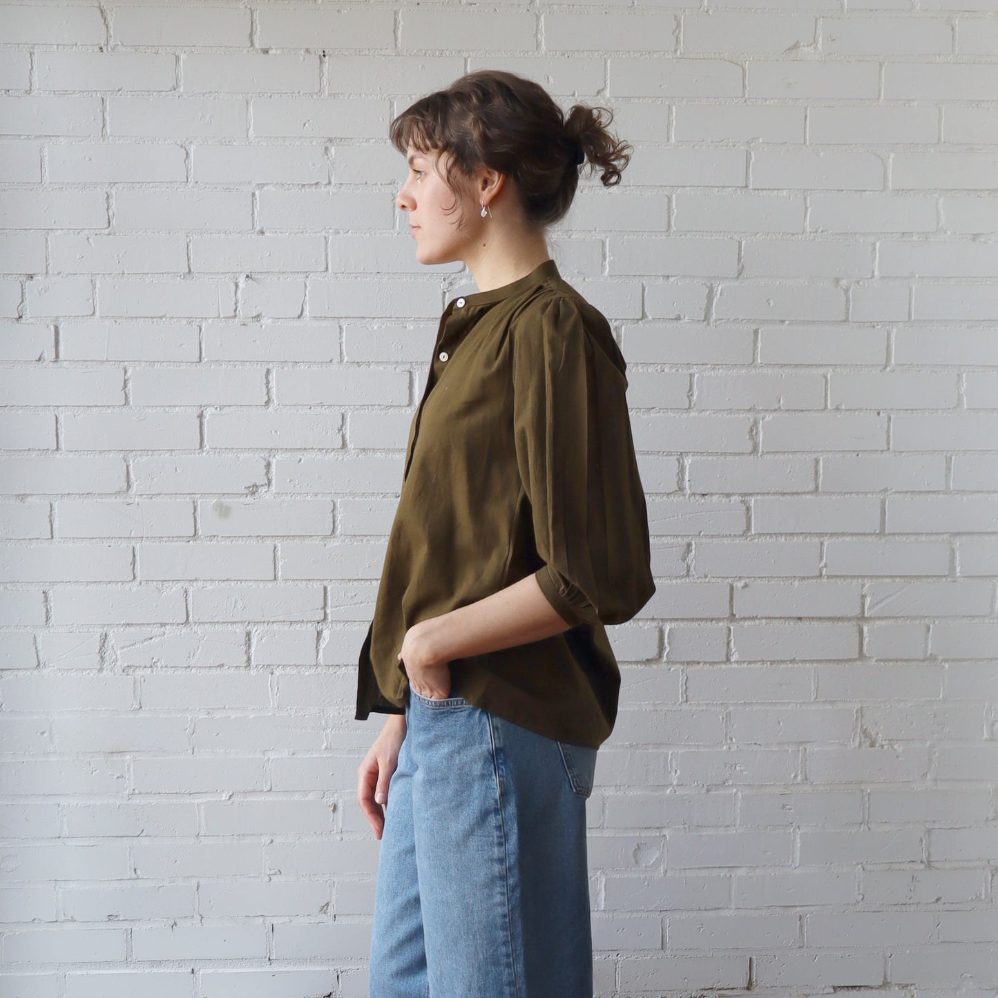 Person wearing an olive green shirt and blue jeans against a white brick wall.