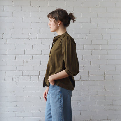 Person wearing an olive green shirt and blue jeans against a white brick wall.