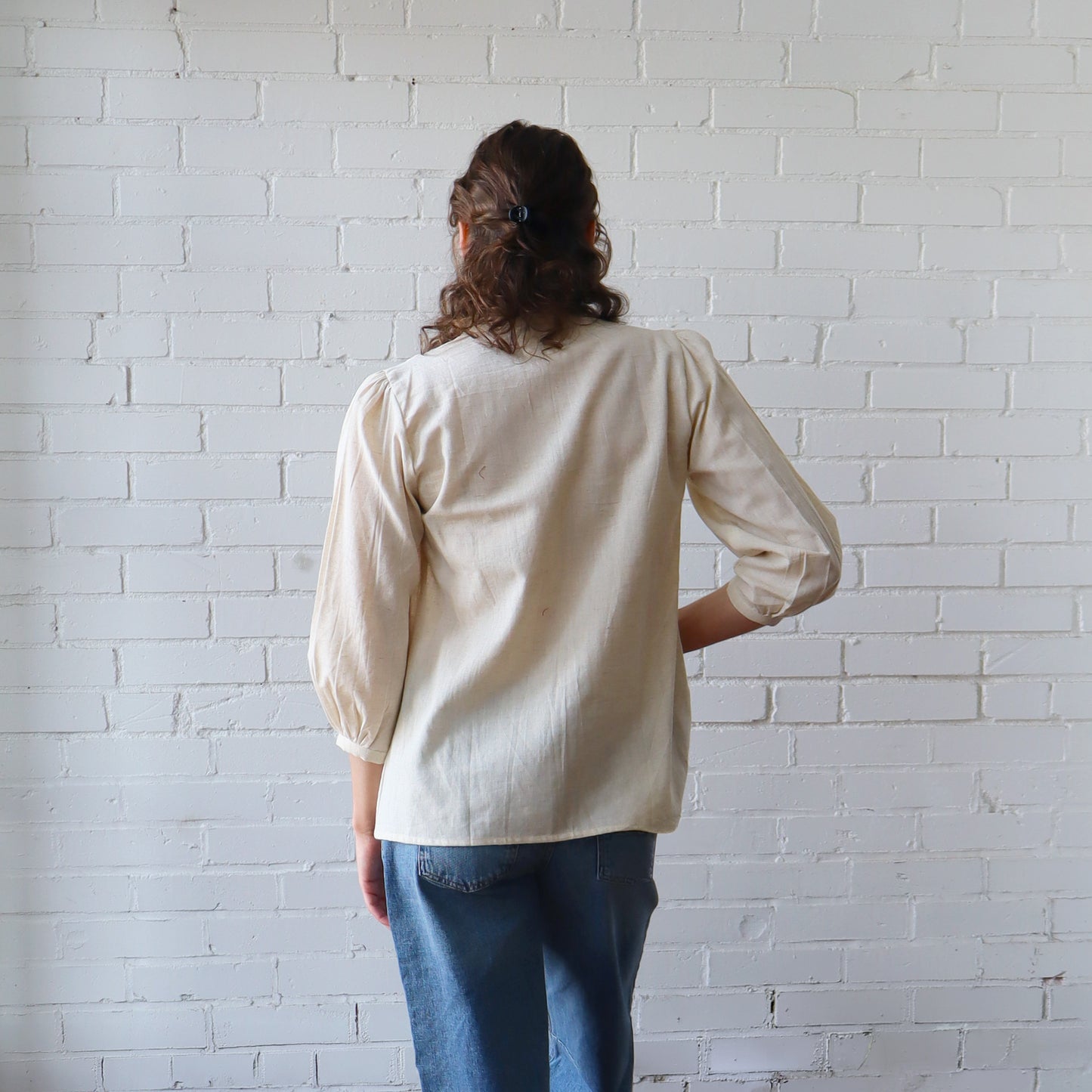 Person wearing a beige blouse and blue jeans against a white brick wall