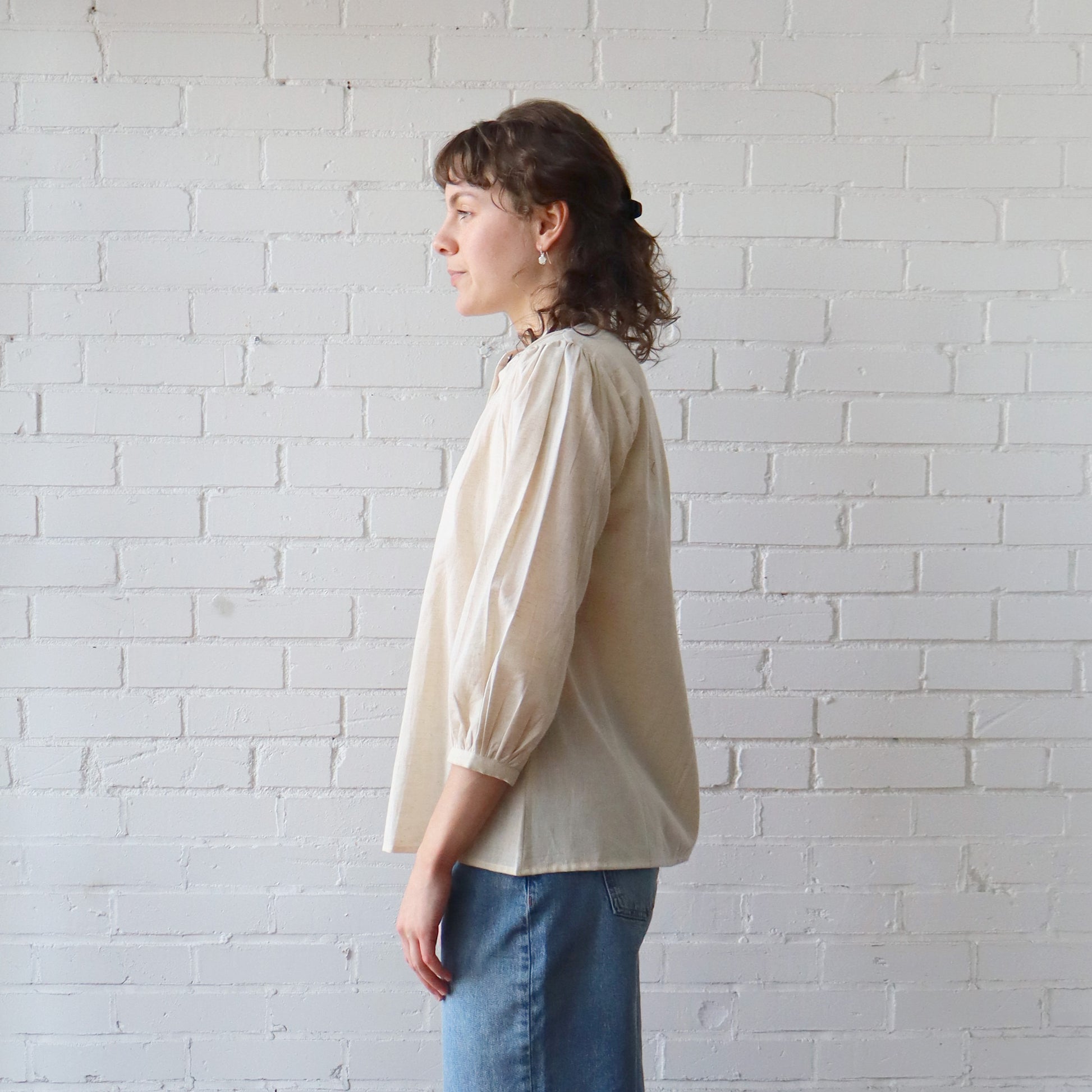 Person wearing a beige blouse and blue jeans against a white brick wall