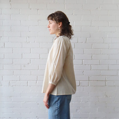 Person wearing a beige blouse and blue jeans against a white brick wall