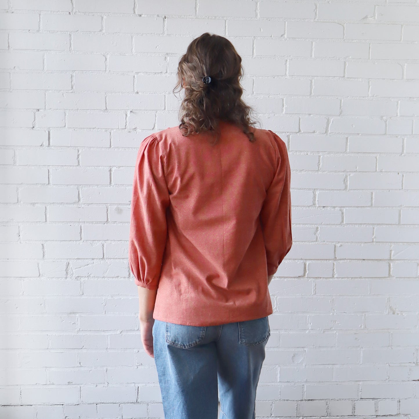 Person wearing a pink blouse and blue jeans standing against a white brick wall.