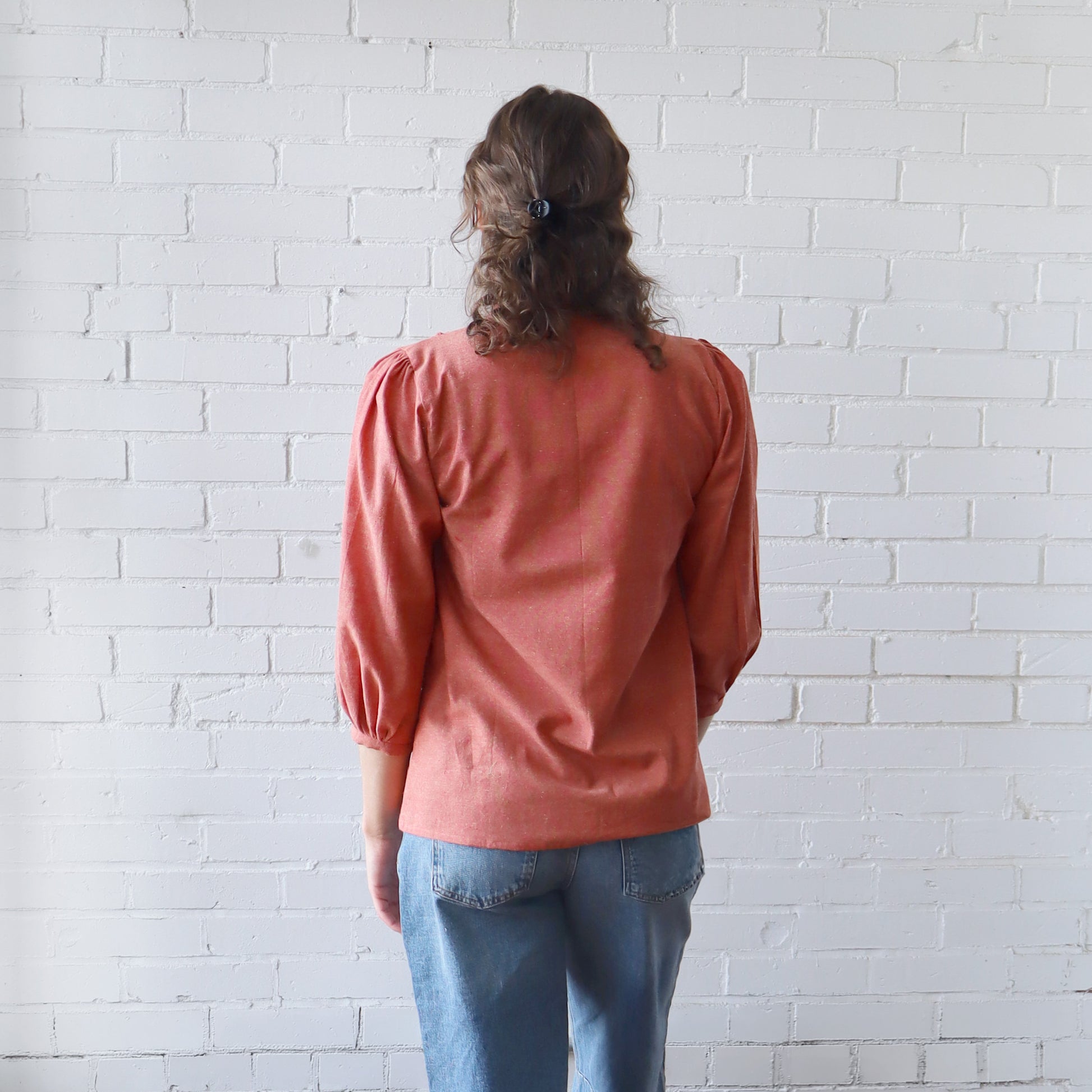 Person wearing a pink blouse and blue jeans standing against a white brick wall.