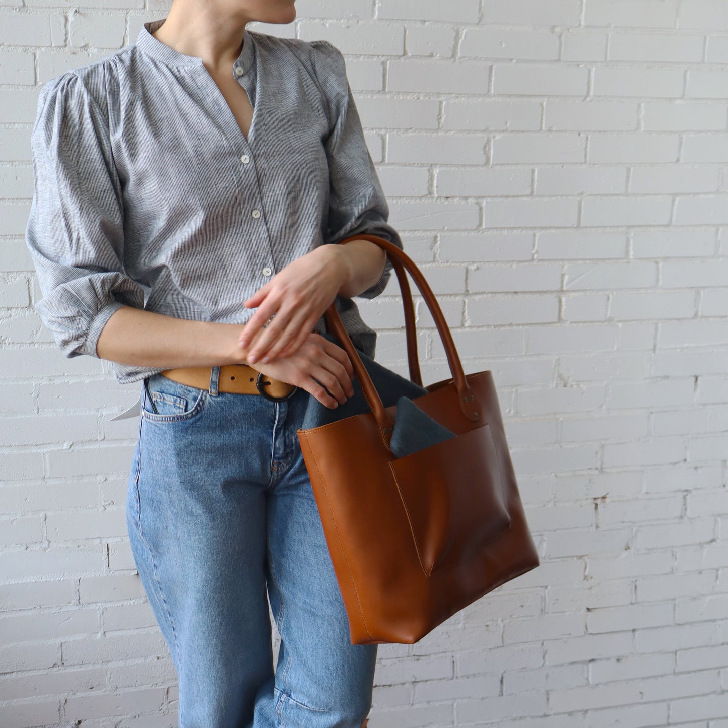 Person holding a brown leather tote bag against a white brick wall.