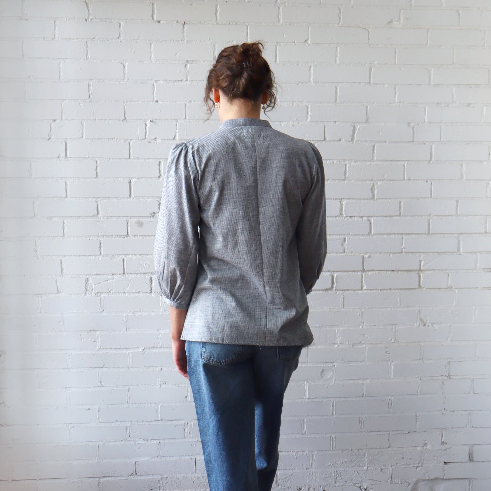 Person wearing a gray shirt and blue jeans standing against a white brick wall.