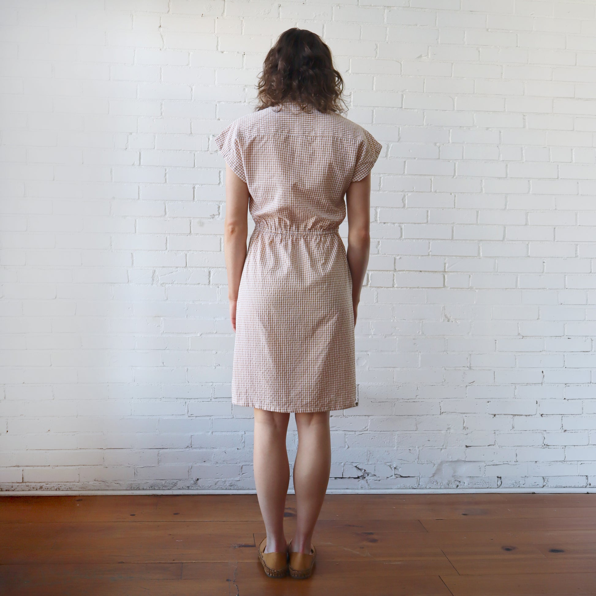 Woman wearing a beige checkered dress standing against a white brick wall.