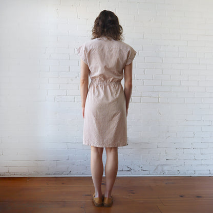 Woman wearing a beige checkered dress standing against a white brick wall.