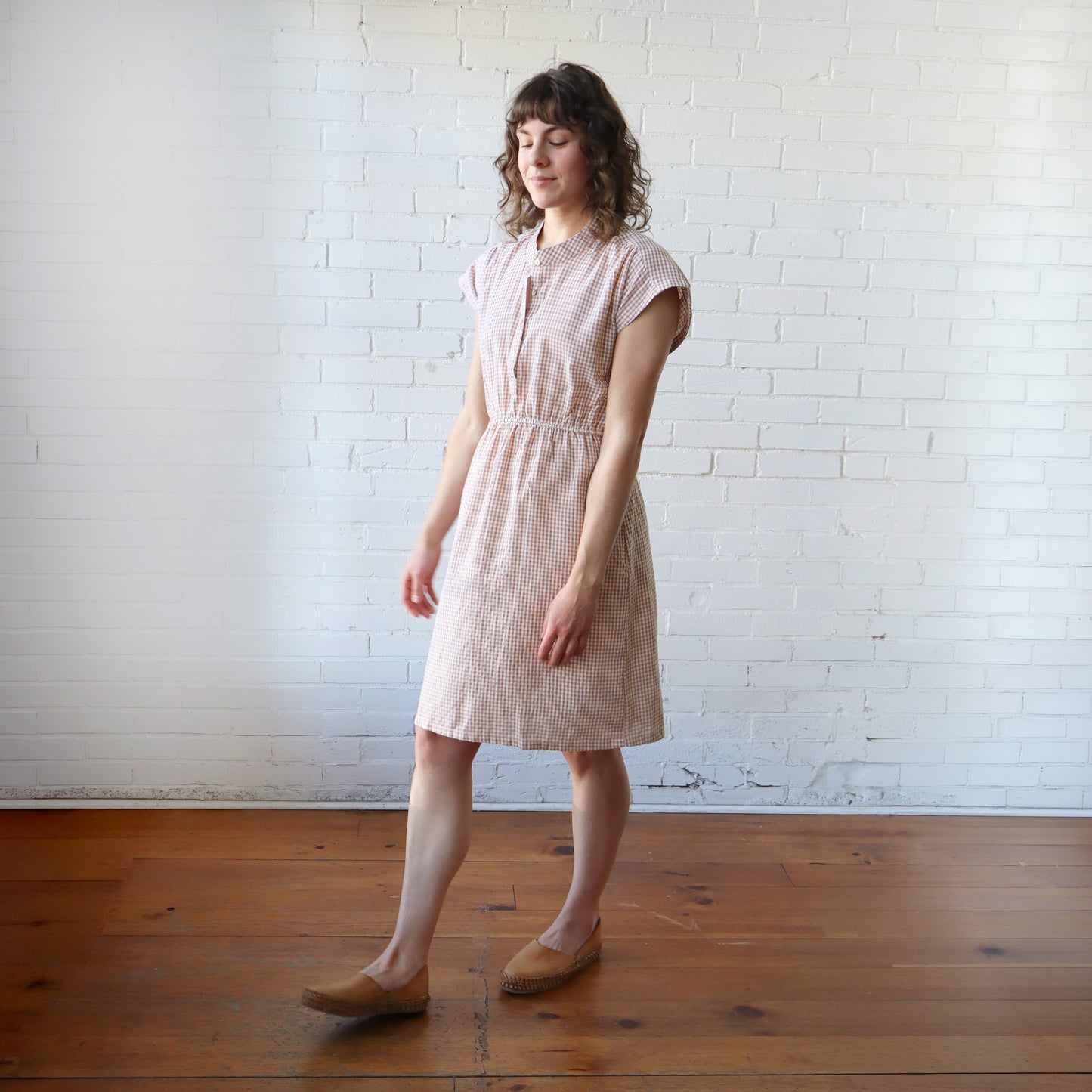 Woman wearing a pink checkered dress standing against a white brick wall.