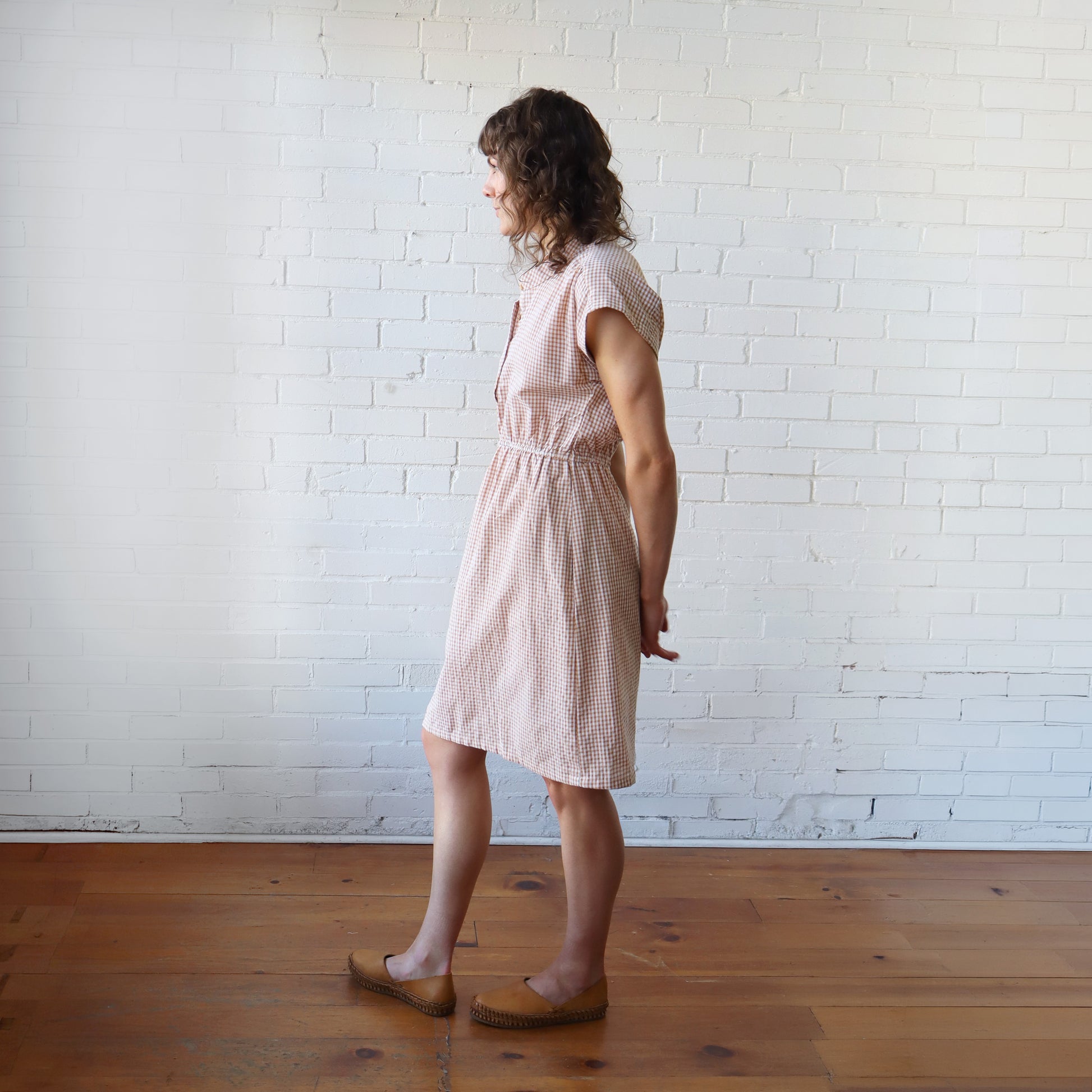 Woman wearing a pink checkered dress standing against a white brick wall.