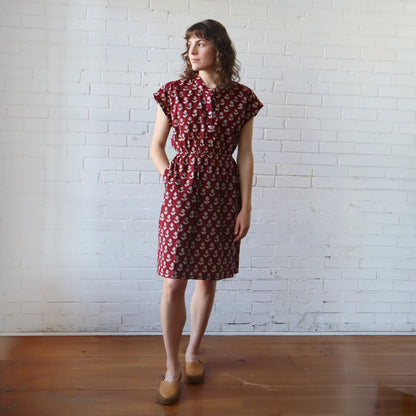 Woman wearing a red patterned dress standing against a white brick wall.