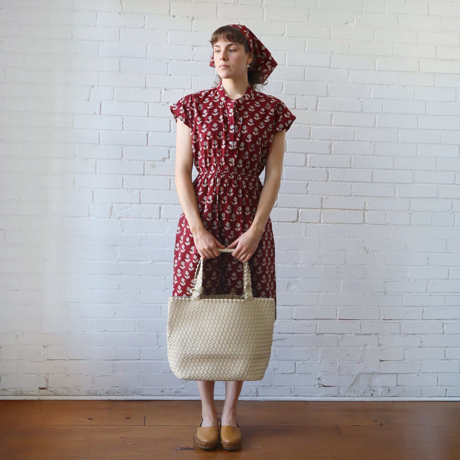 Woman in a red patterned dress holding a woven bag against a white brick wall.