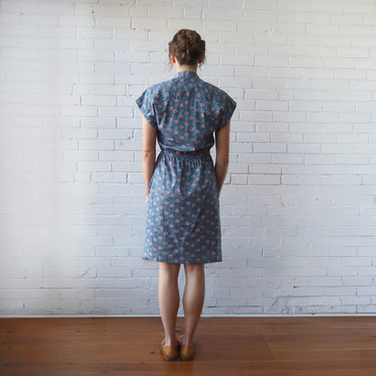 Woman wearing a blue patterned dress standing against a white brick wall.