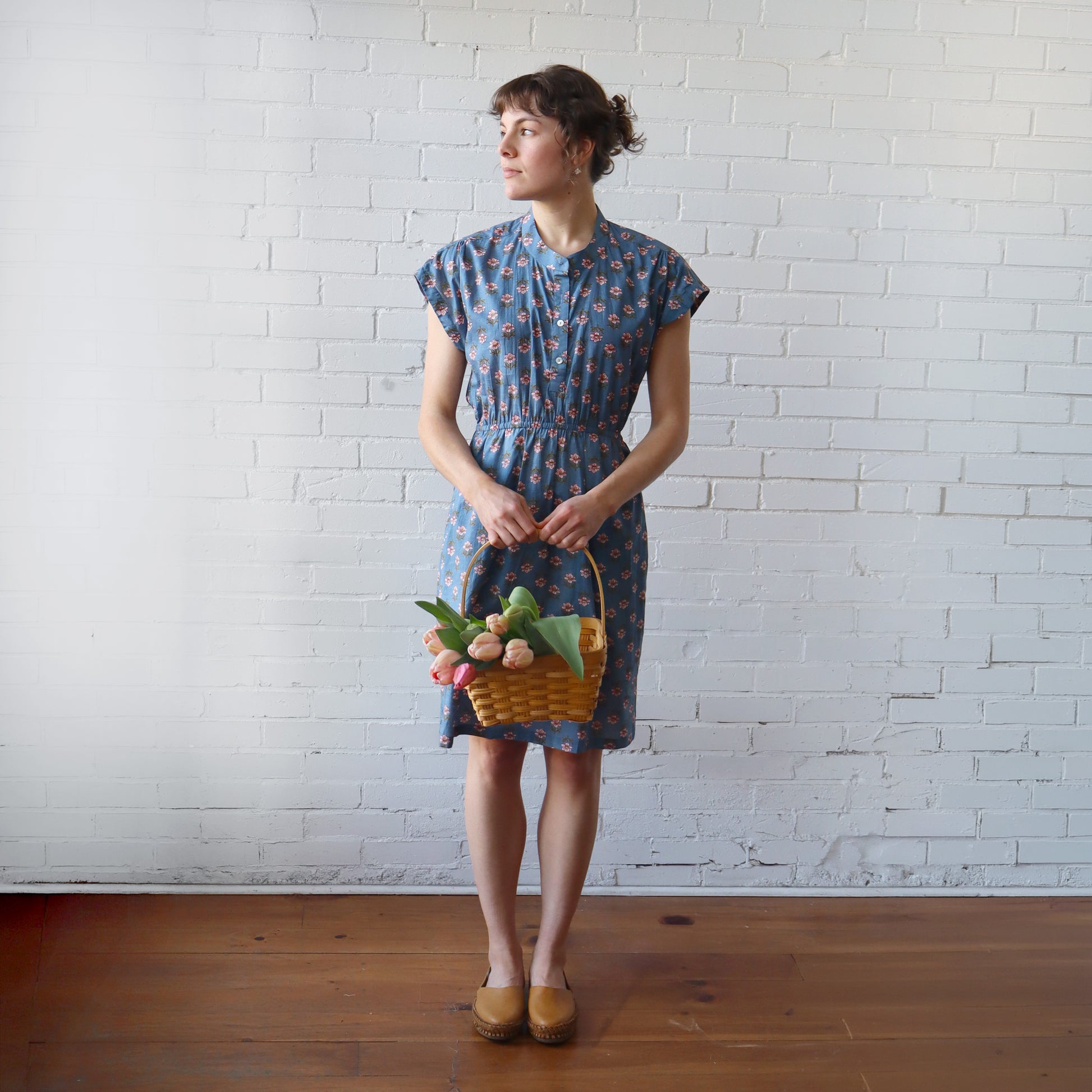 Woman in a blue patterned dress holding a basket of flowers against a white brick wall.