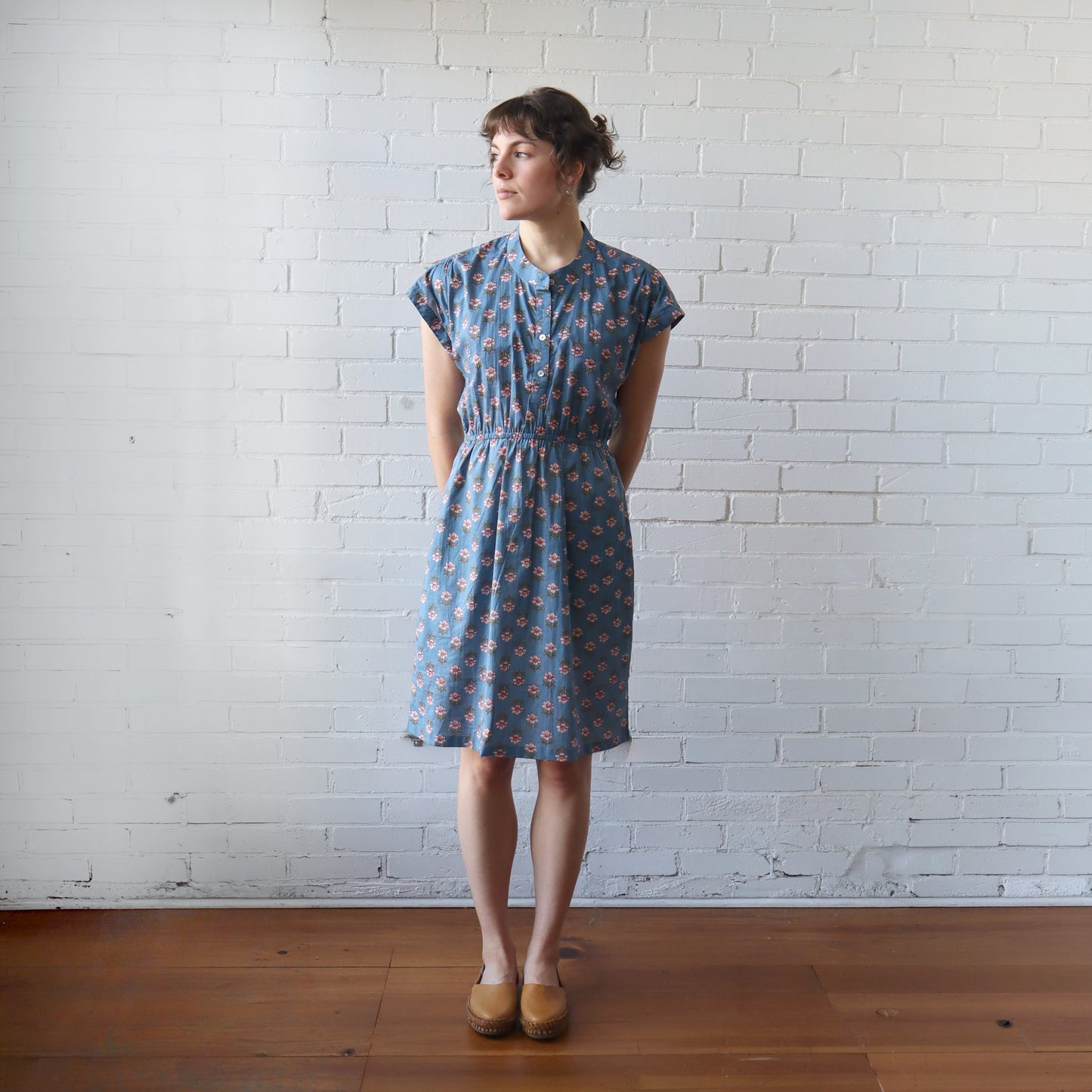 Woman wearing a blue floral dress standing against a white brick wall.