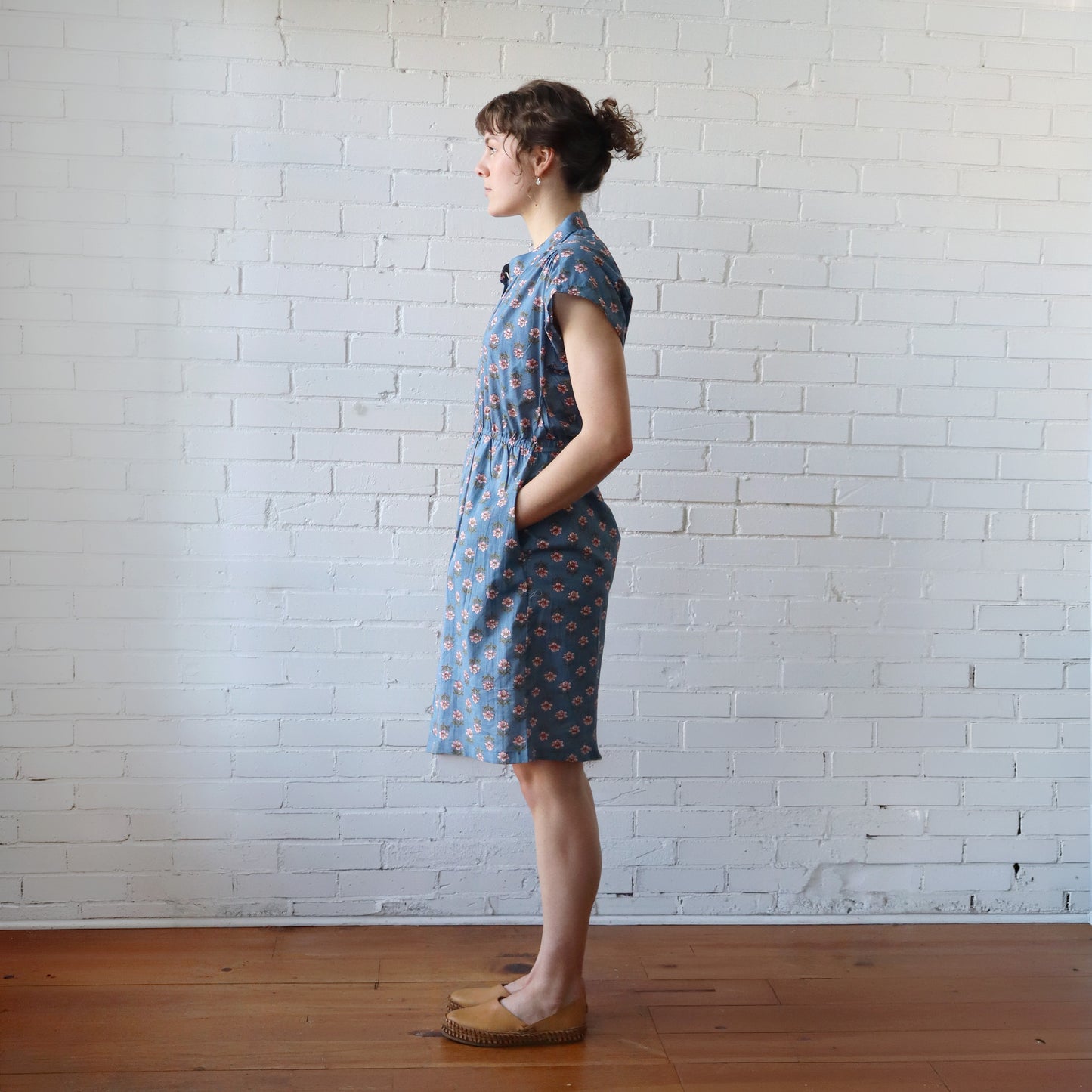 Woman wearing a blue floral dress standing against a white brick wall.