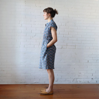 Woman wearing a blue floral dress standing against a white brick wall.