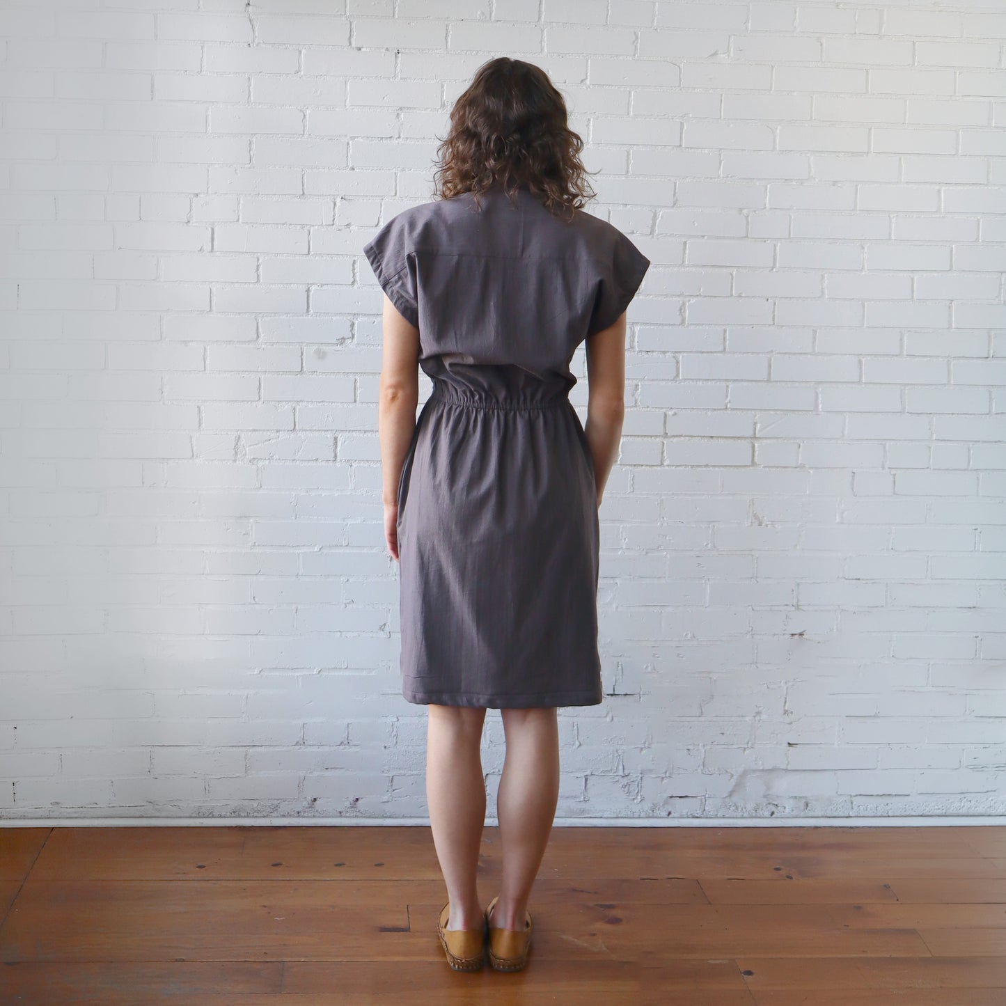 Woman wearing a gray dress standing against a white brick wall.