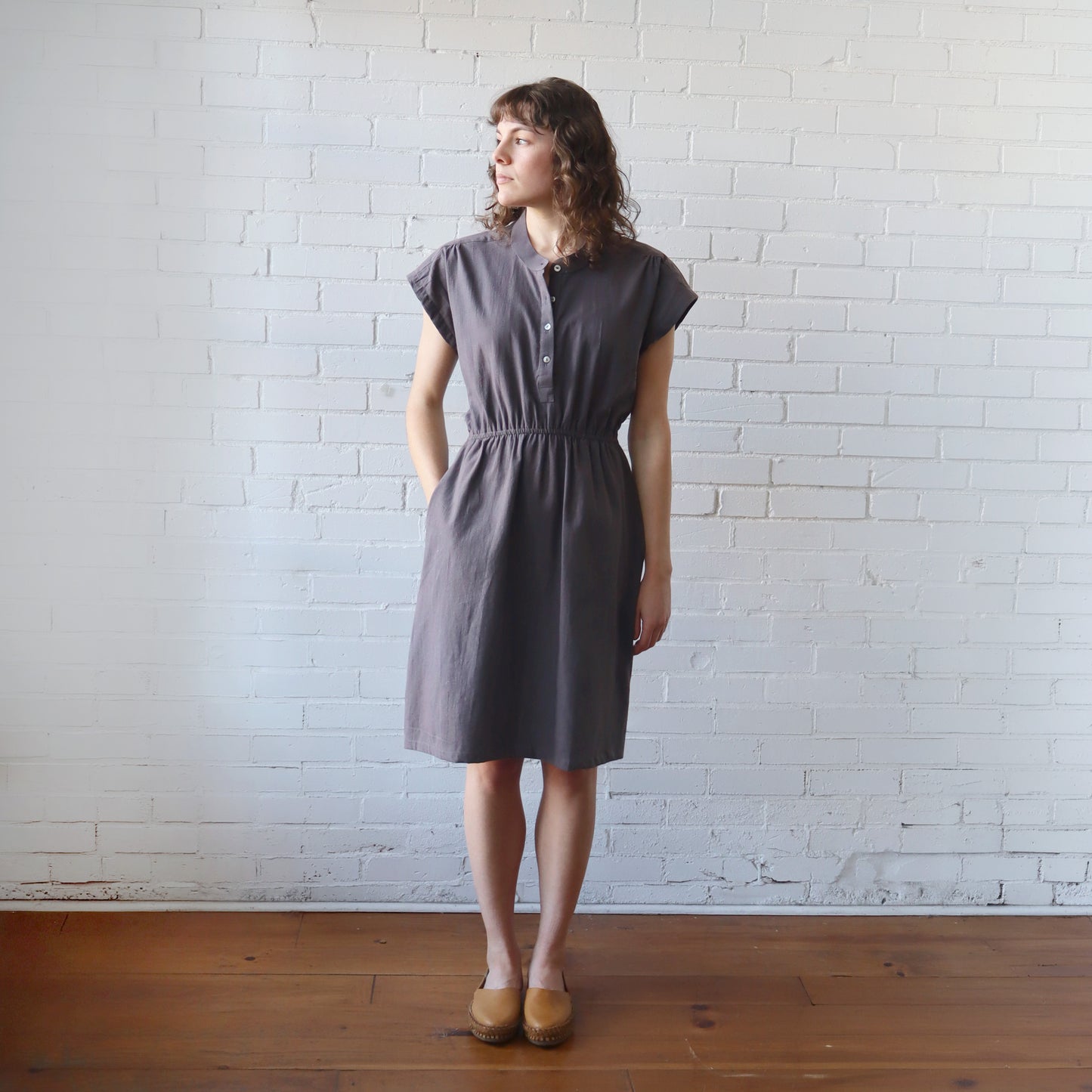 Woman wearing a gray dress standing against a white brick wall.