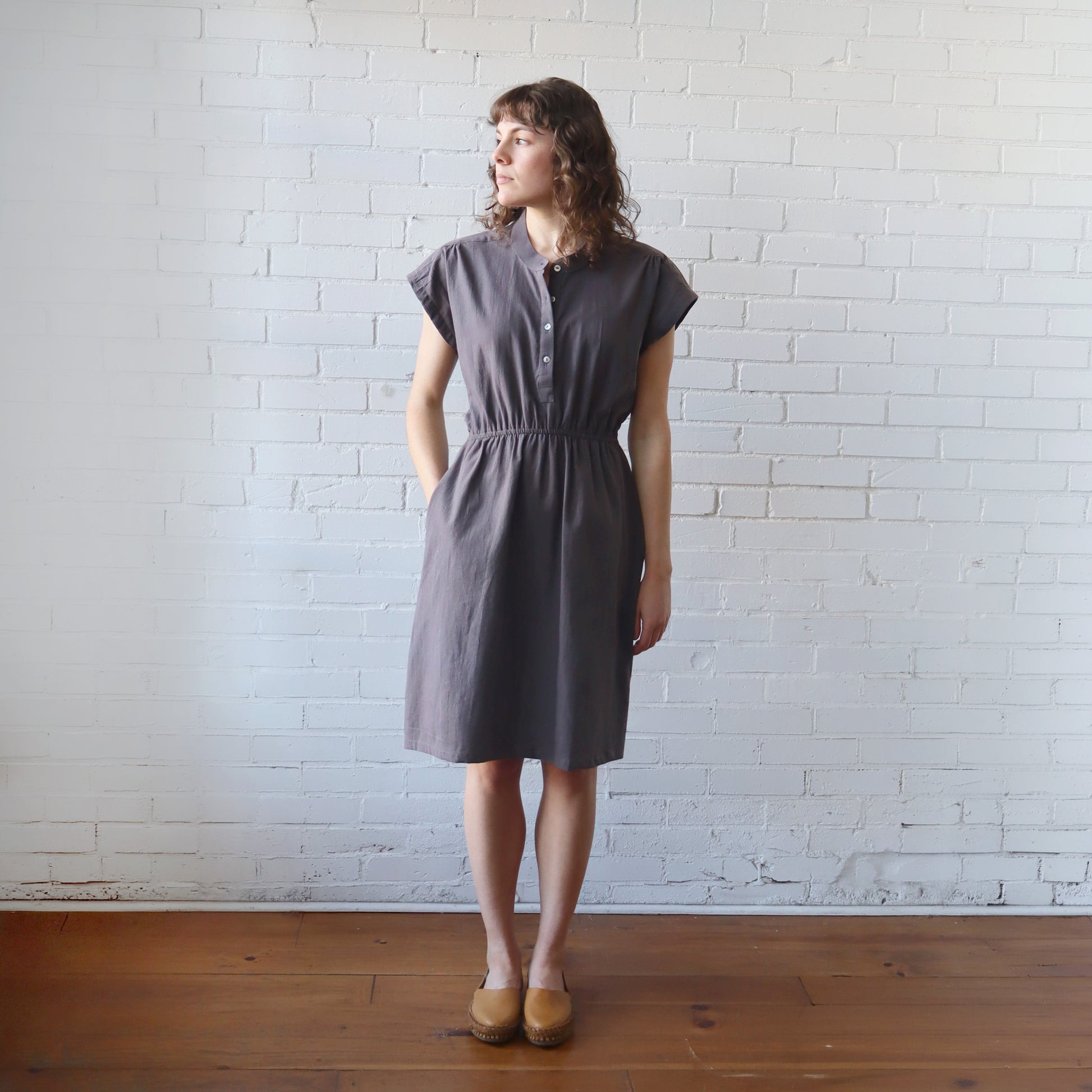 Woman wearing a gray dress standing against a white brick wall.