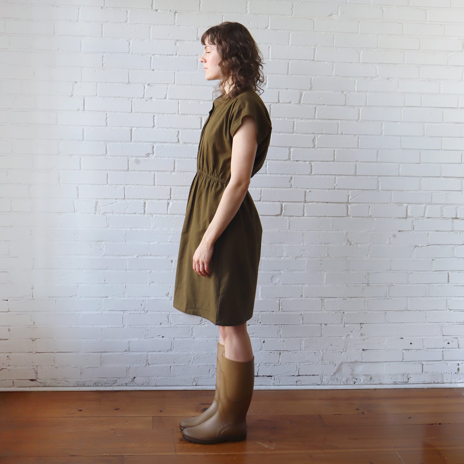 Woman wearing a green dress and boots standing against a white brick wall.