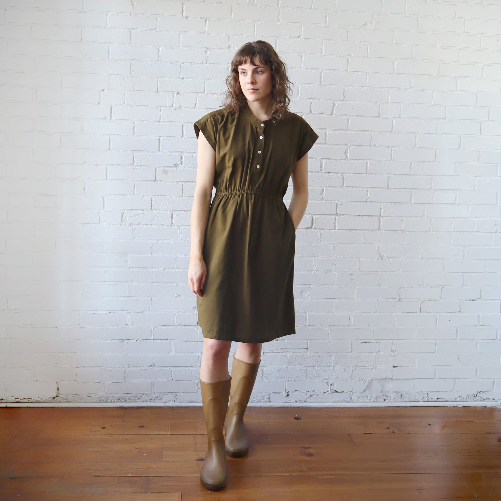 Woman wearing a green dress and brown boots standing against a white brick wall.