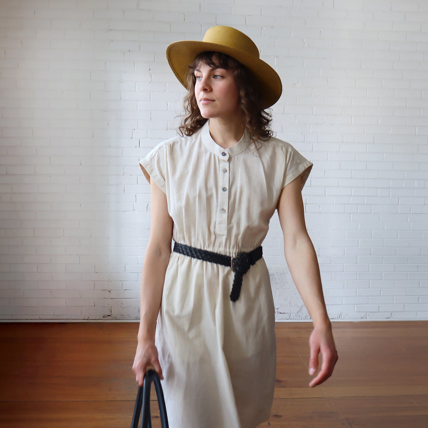 Woman wearing a white dress and yellow hat against a white brick wall.