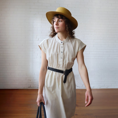 Woman wearing a white dress and yellow hat against a white brick wall.
