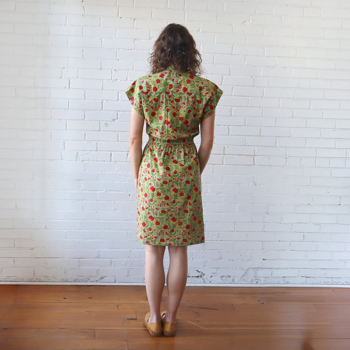 Woman wearing a green floral dress standing against a white brick wall.