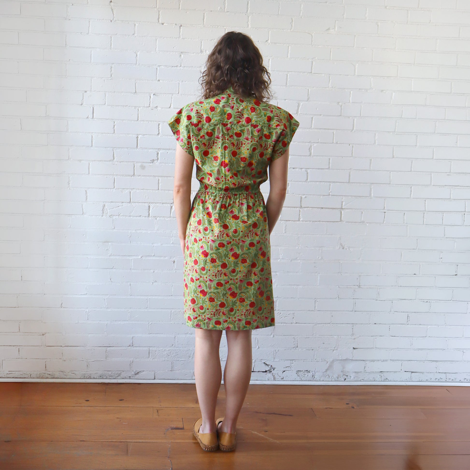 Woman wearing a green floral dress standing against a white brick wall.