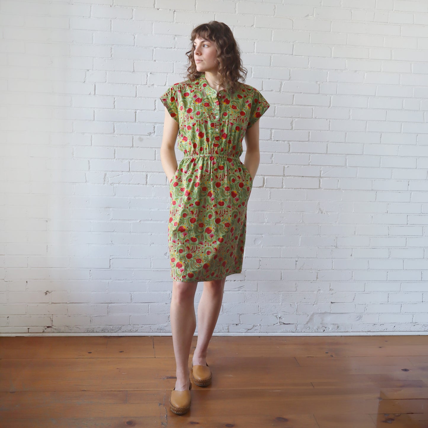 Woman wearing a green floral dress standing against a white brick wall.
