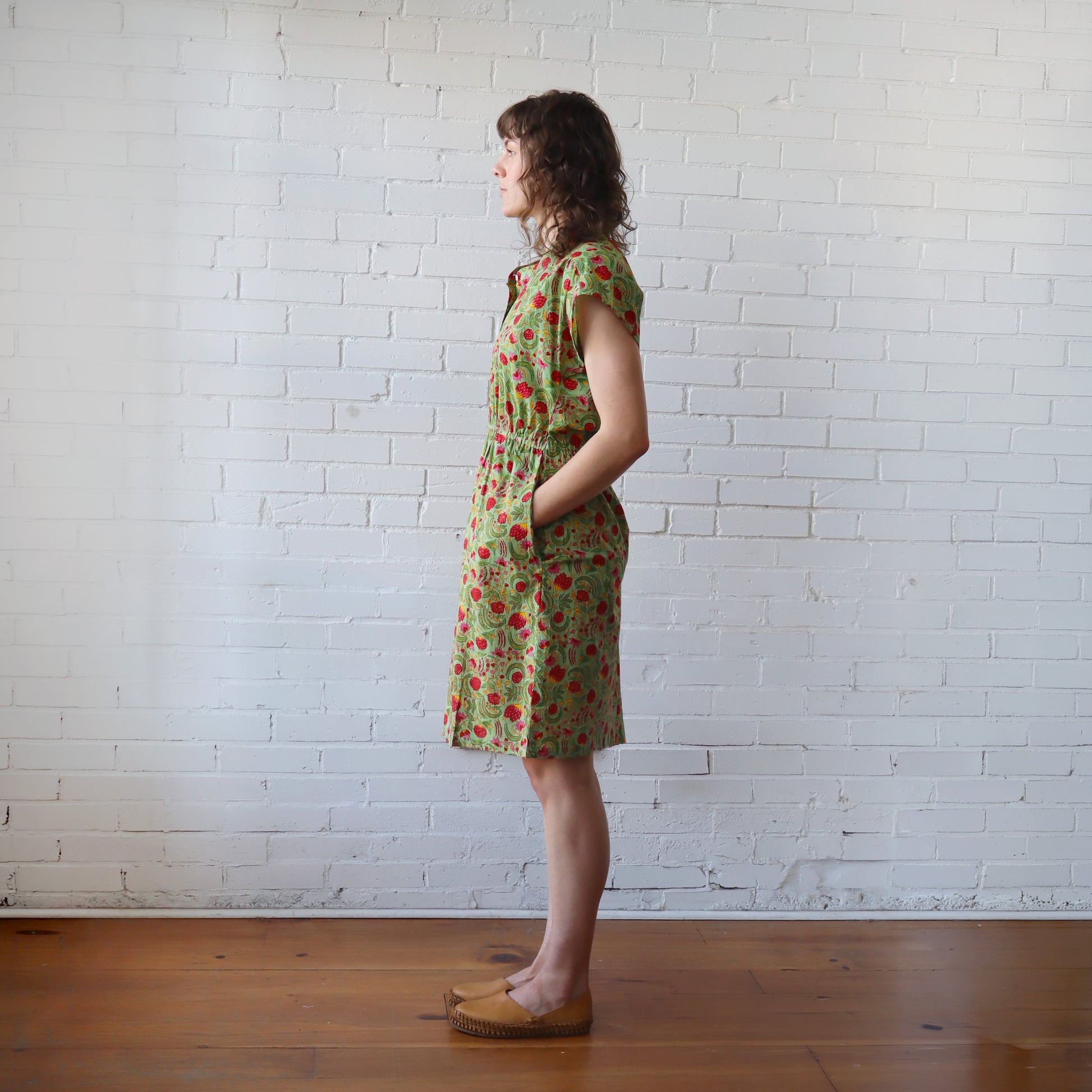 Woman wearing a green floral dress standing against a white brick wall.