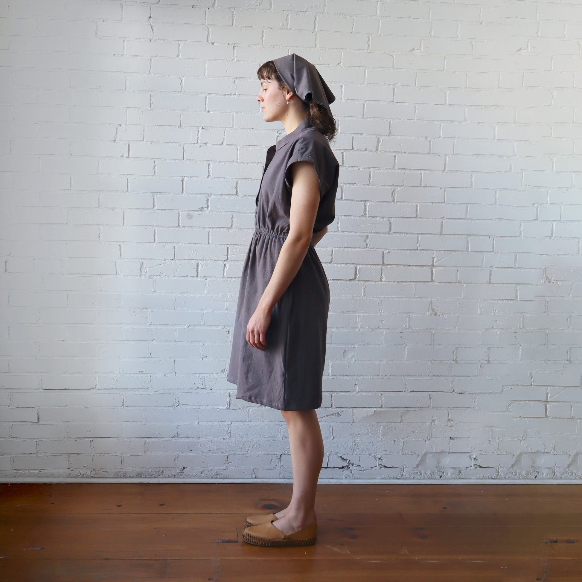 Woman wearing a gray dress and headscarf standing against a white brick wall.