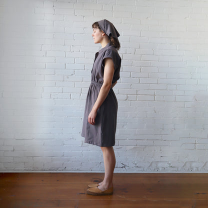 Woman wearing a gray dress and headscarf standing against a white brick wall.
