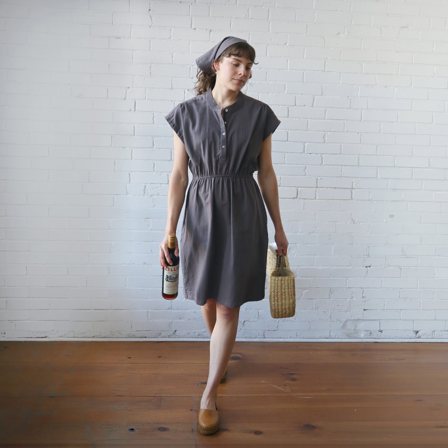 Woman in a gray dress holding a bottle and a woven bag against a white brick wall.