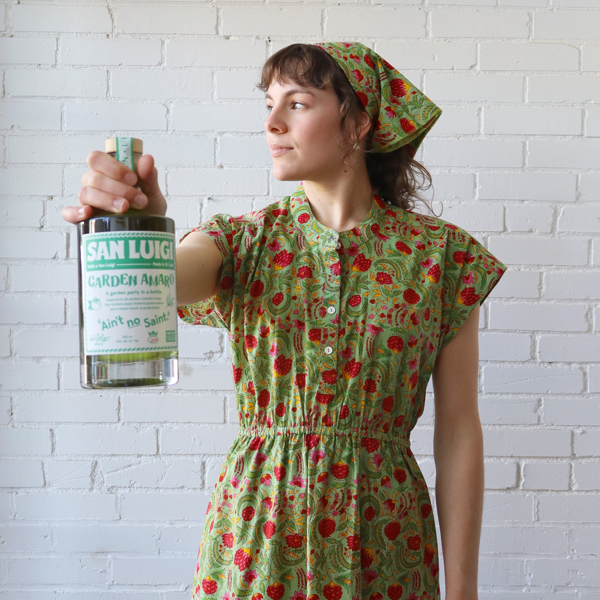 Woman in a green floral dress holding a bottle against a white brick wall