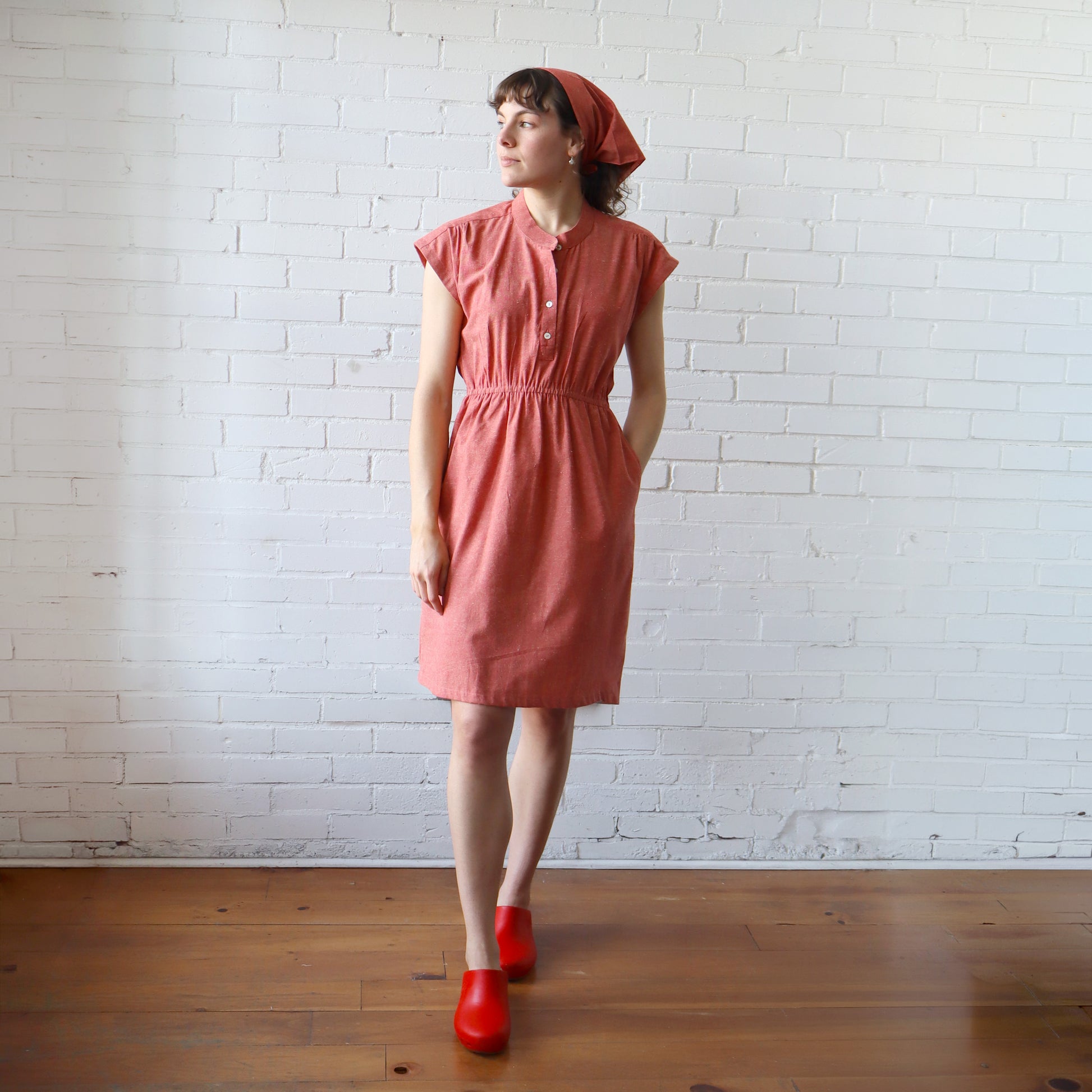 Woman wearing a pink dress and red shoes against a white brick wall.