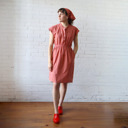 Woman wearing a pink dress and red shoes against a white brick wall.