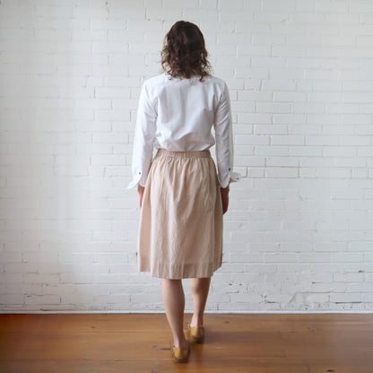Person wearing a white shirt and beige checkered skirt standing against a white brick wall.