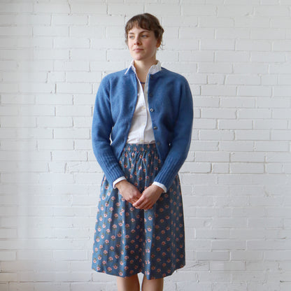 Woman wearing a blue cardigan and patterned skirt against a white brick wall.