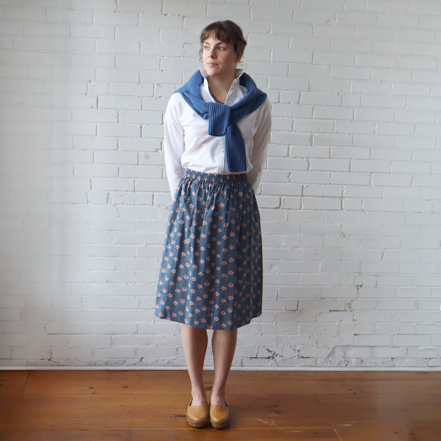 Woman wearing a white blouse, blue patterned skirt, and blue scarf against a white brick wall.
