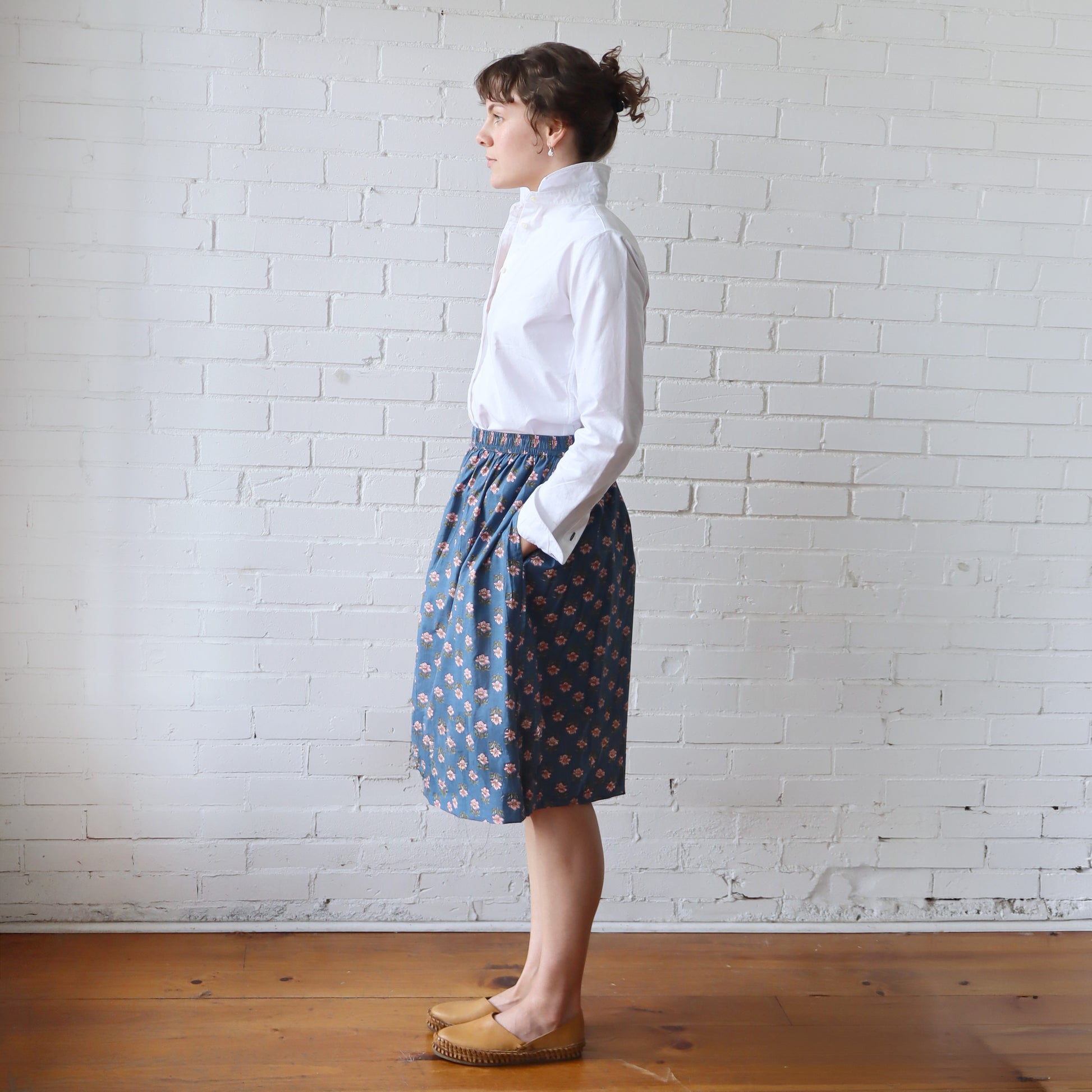 Woman wearing a white blouse and blue patterned skirt against a white brick wall.