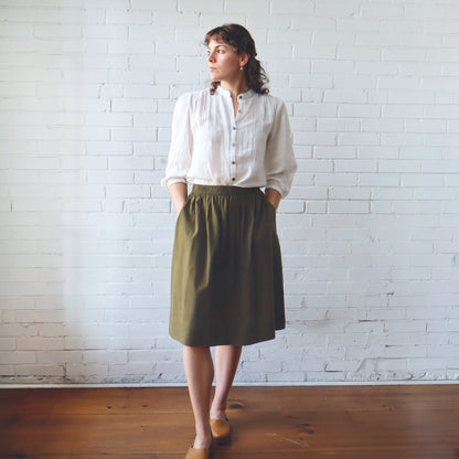 Woman wearing a white shirt and green skirt standing against a white brick wall.