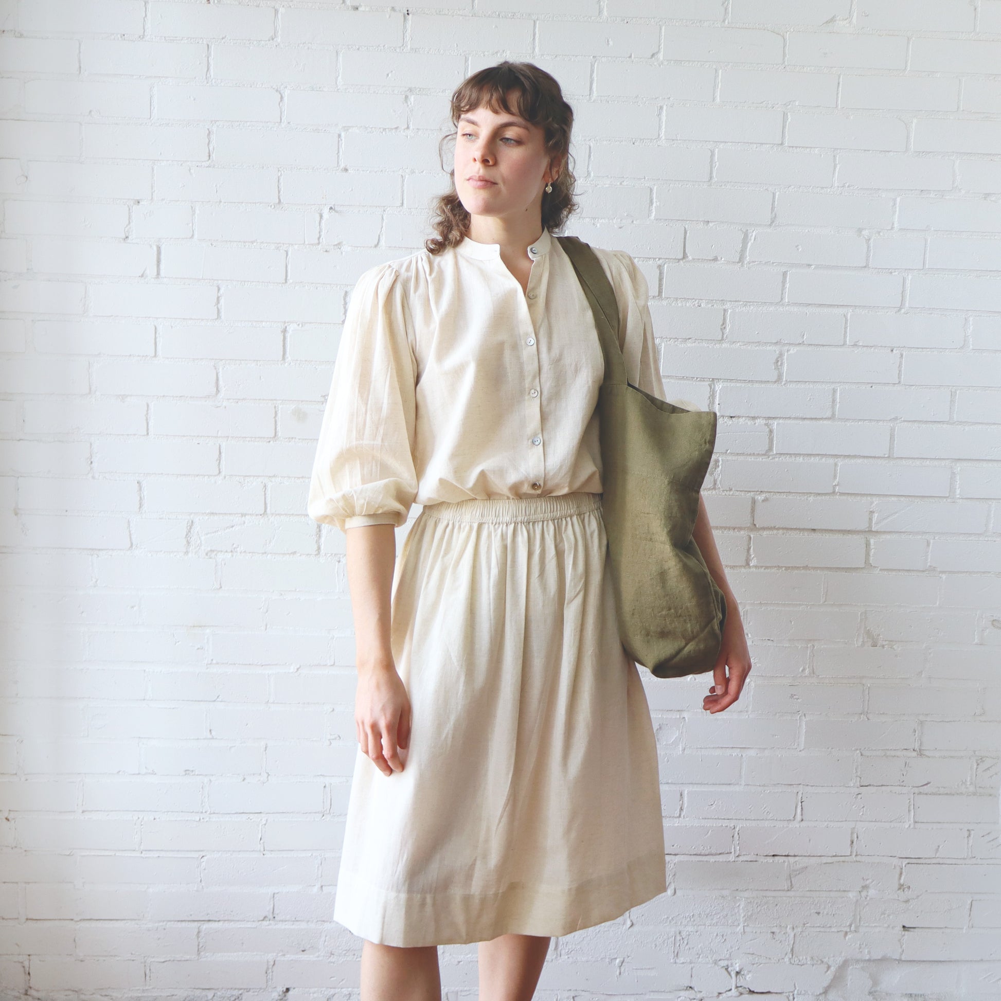 Woman in a beige blouse and skirt holding a green bag against a white brick wall
