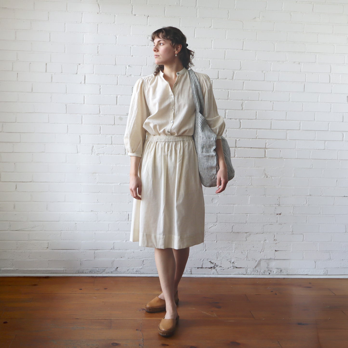 Woman in a beige blouse and skirt holding a striped bag against a white brick wall.