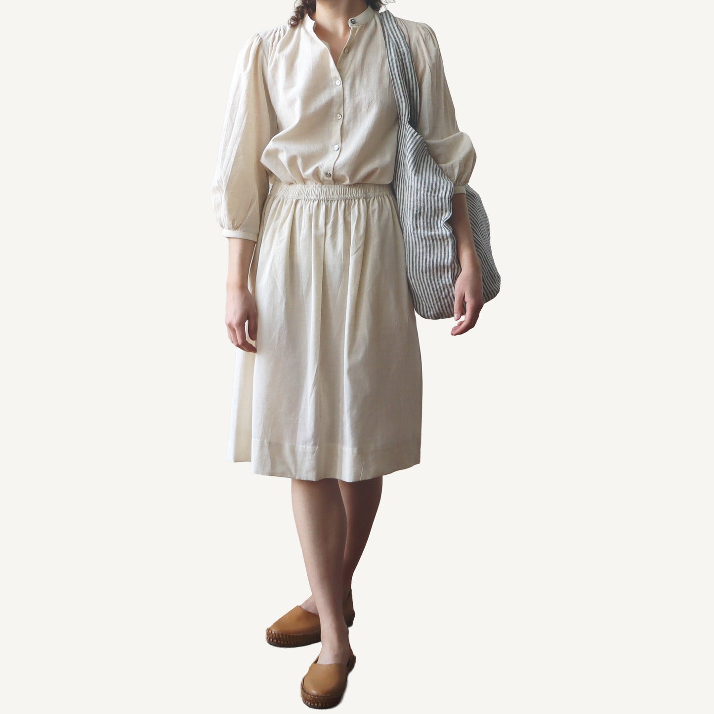 Person wearing a beige blouse and skirt with a striped bag on a plain background