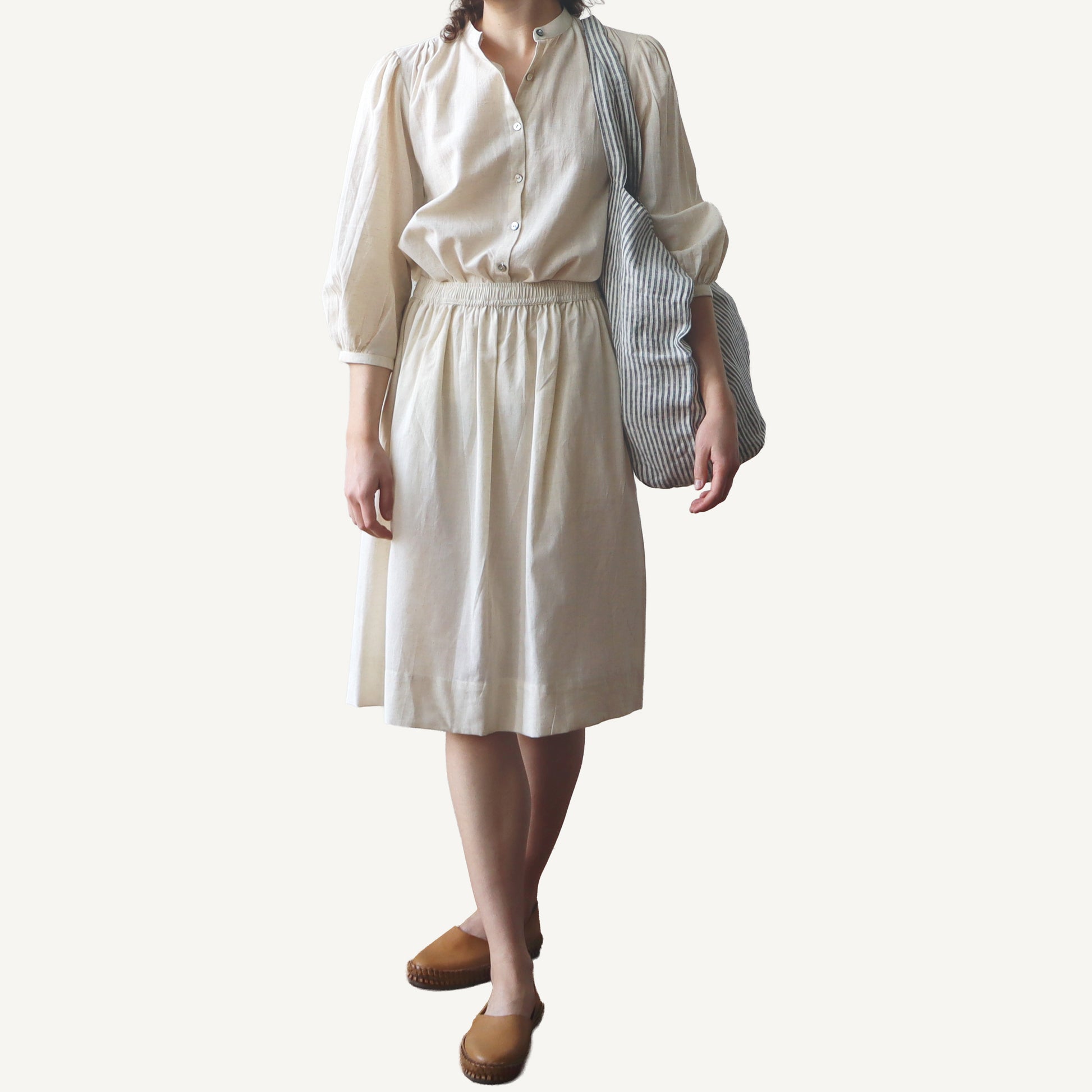 Person wearing a beige blouse and skirt with a striped bag on a plain background