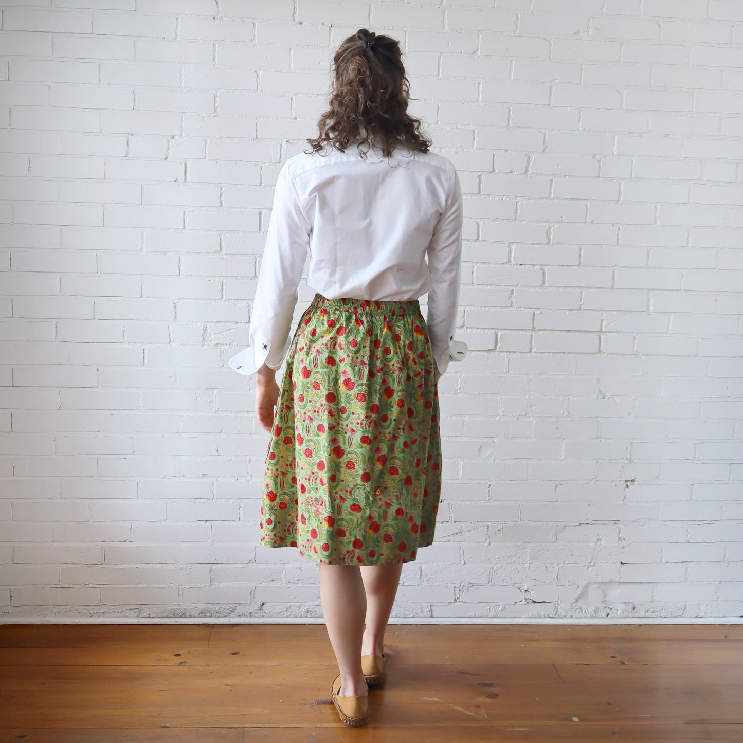 Person wearing a white shirt and green floral skirt against a white brick wall.