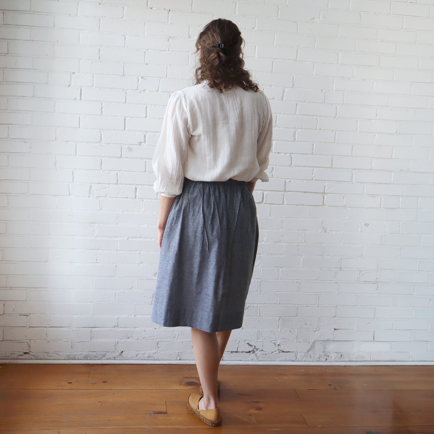 Person wearing a white blouse and gray skirt standing against a white brick wall.