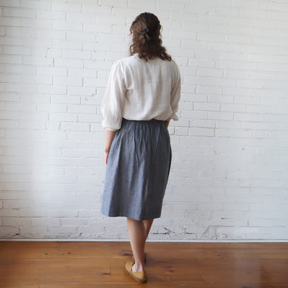 Person wearing a white blouse and gray skirt standing against a white brick wall.